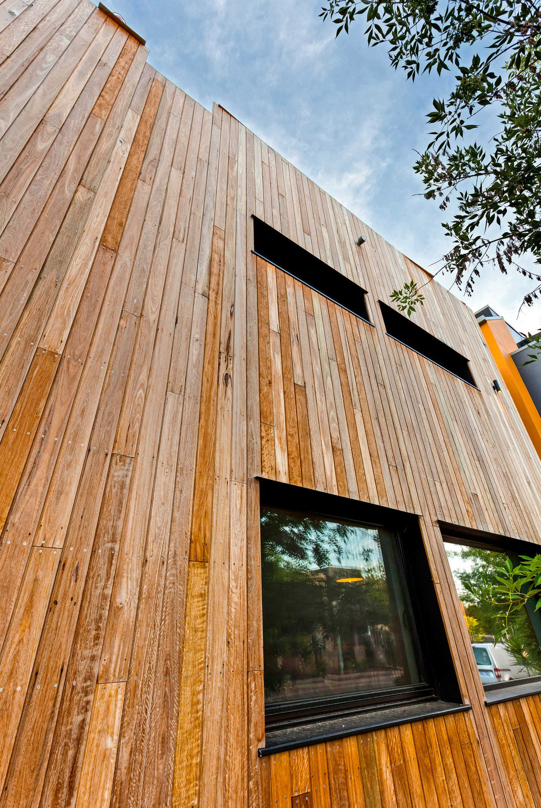 Timber-clad facade with deep-set windows under trees and blue sky